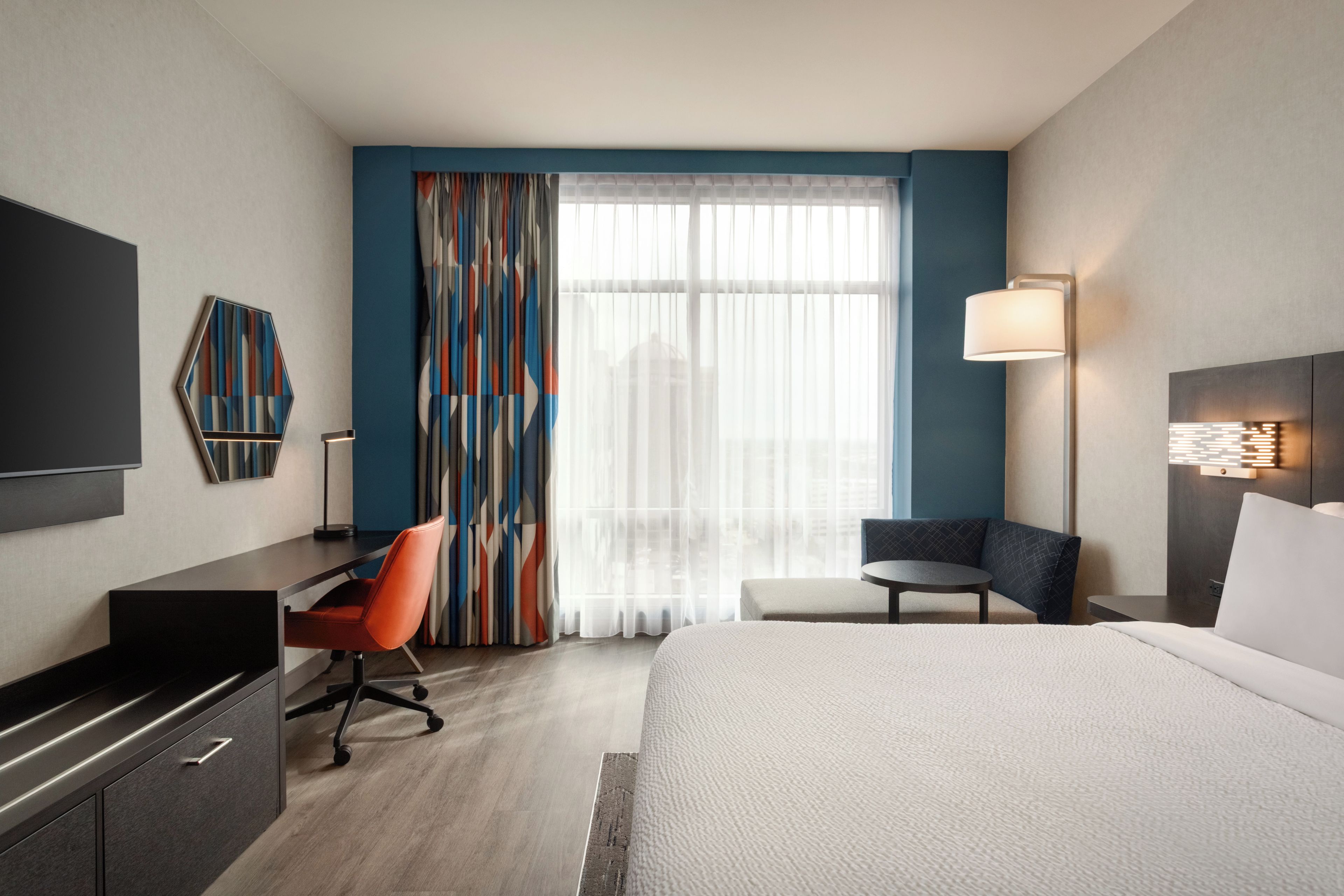 A hotel room with a bed, a desk, a chair, a TV, and a window with curtains.