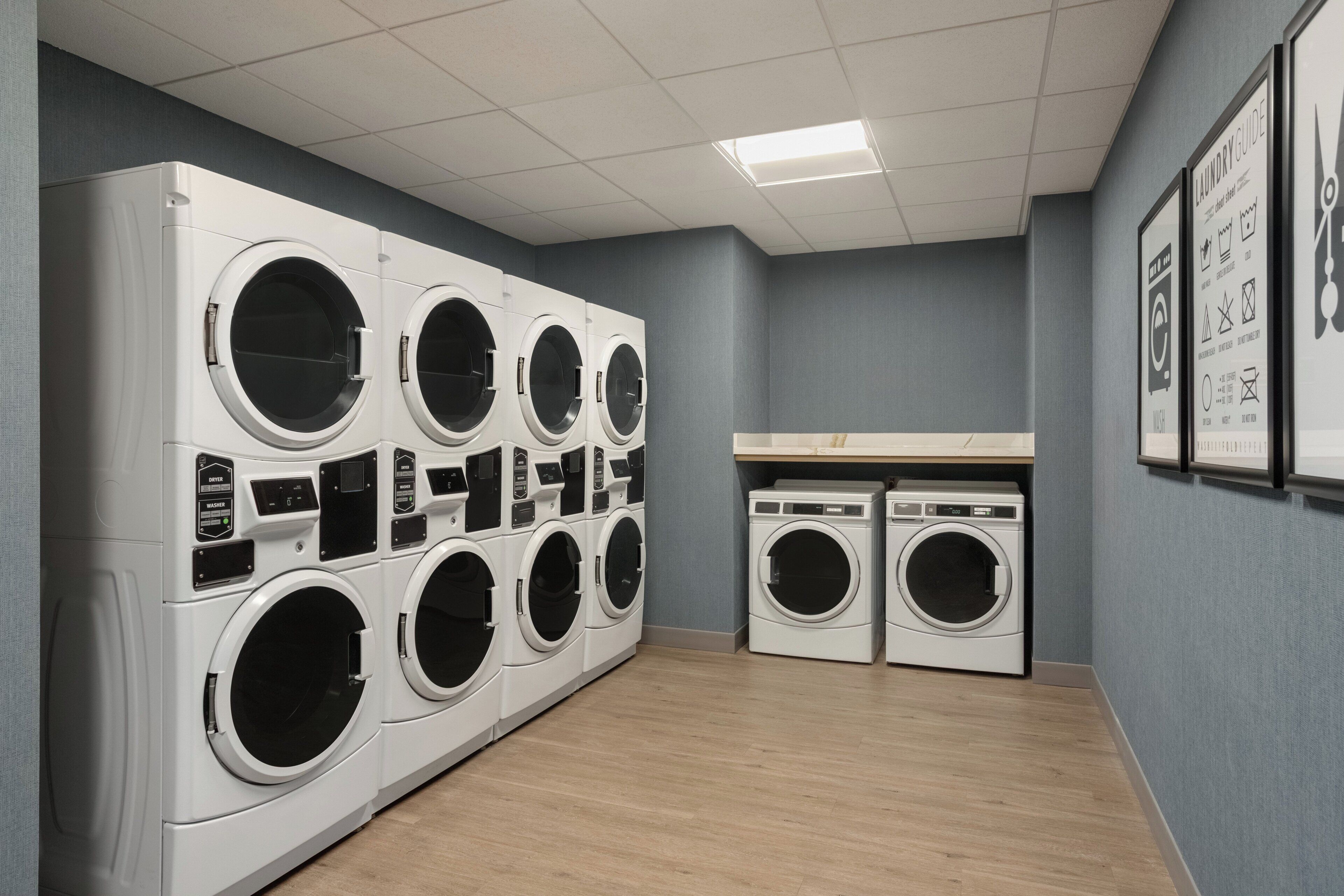 A laundry facility with multiple washing machines and dryers, a tiled ceiling, and a grey wall with a laundry guide.
