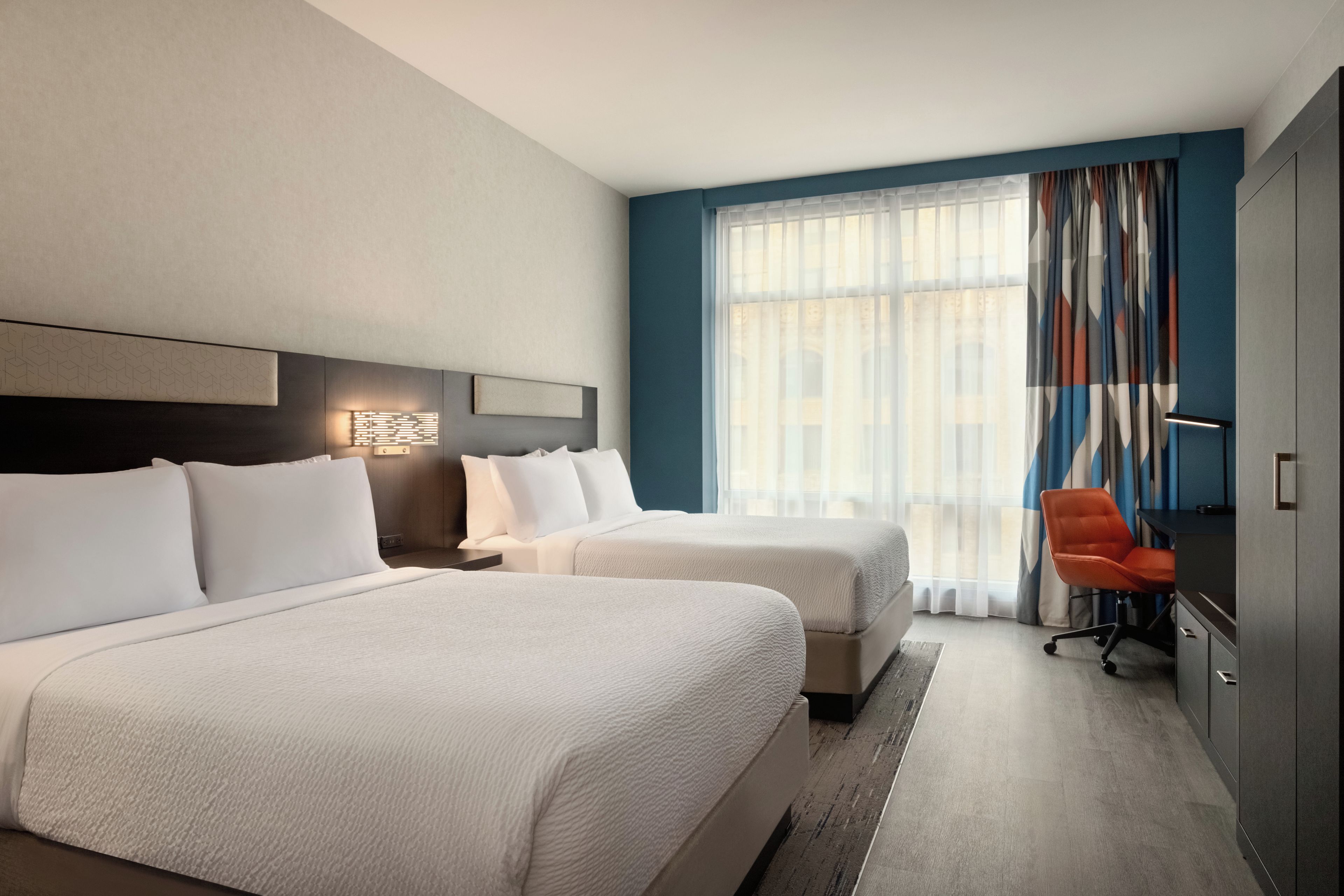 Habitación de hotel con dos camas, un escritorio, una silla y un ventanal con cortinas.
