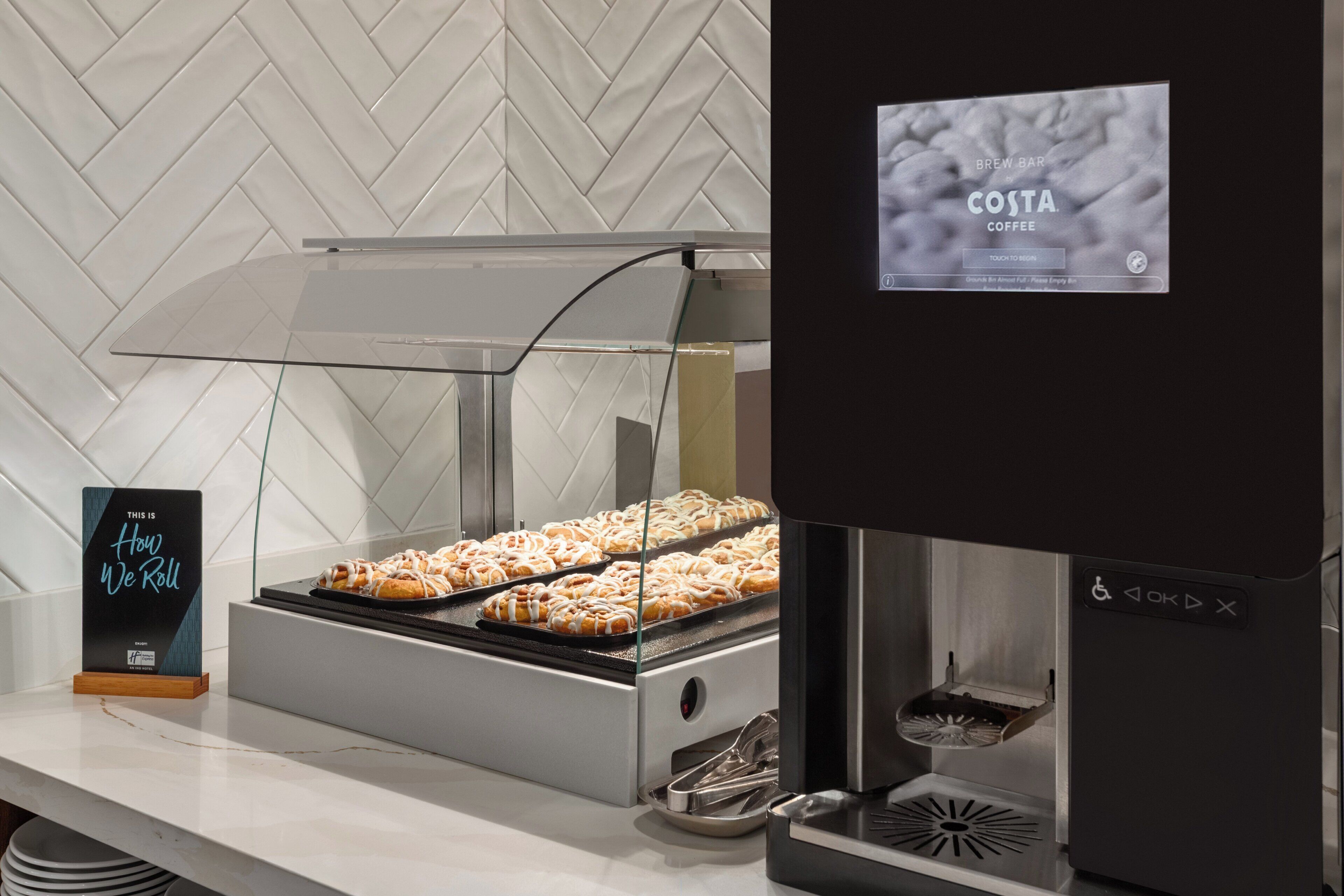 A coffee station with a sign that reads 'BREW BAR by COSTA COFFEE' and a sign that says 'THIS IS How We Roll.'