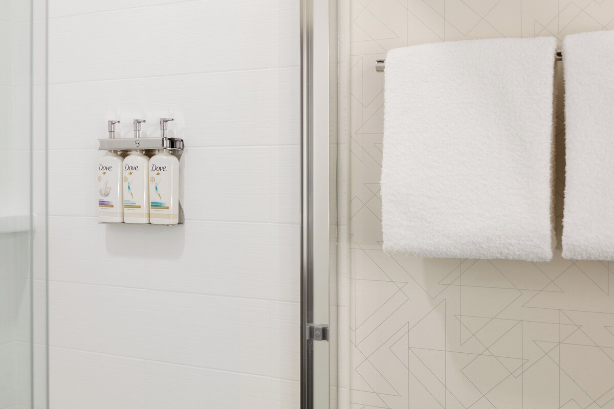 A bathroom with white tiles, a glass shower door, and three Dove shampoo bottles on a wall-mounted shelf.