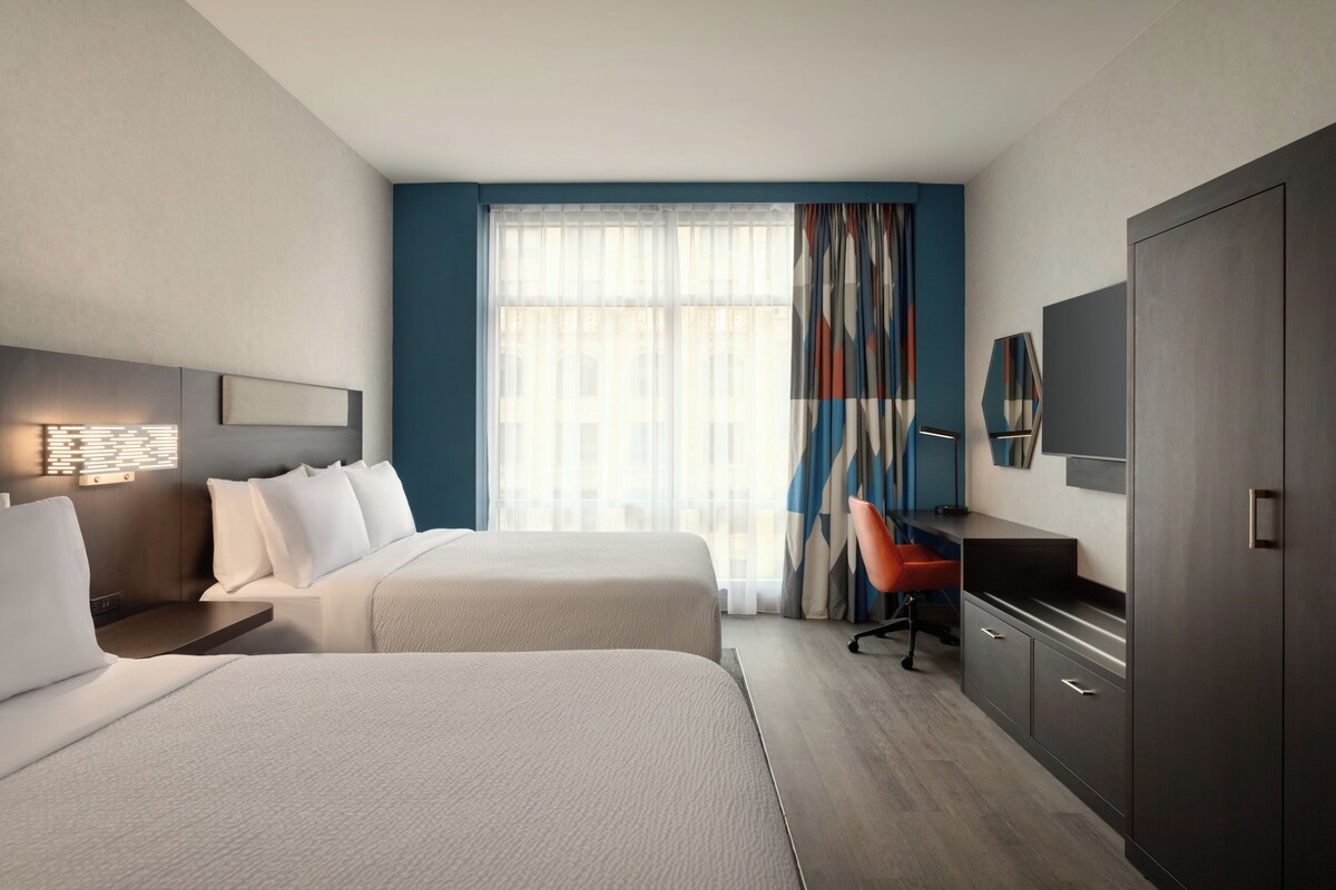A hotel room with two beds, a desk, a chair, a wardrobe, and a large window with curtains.