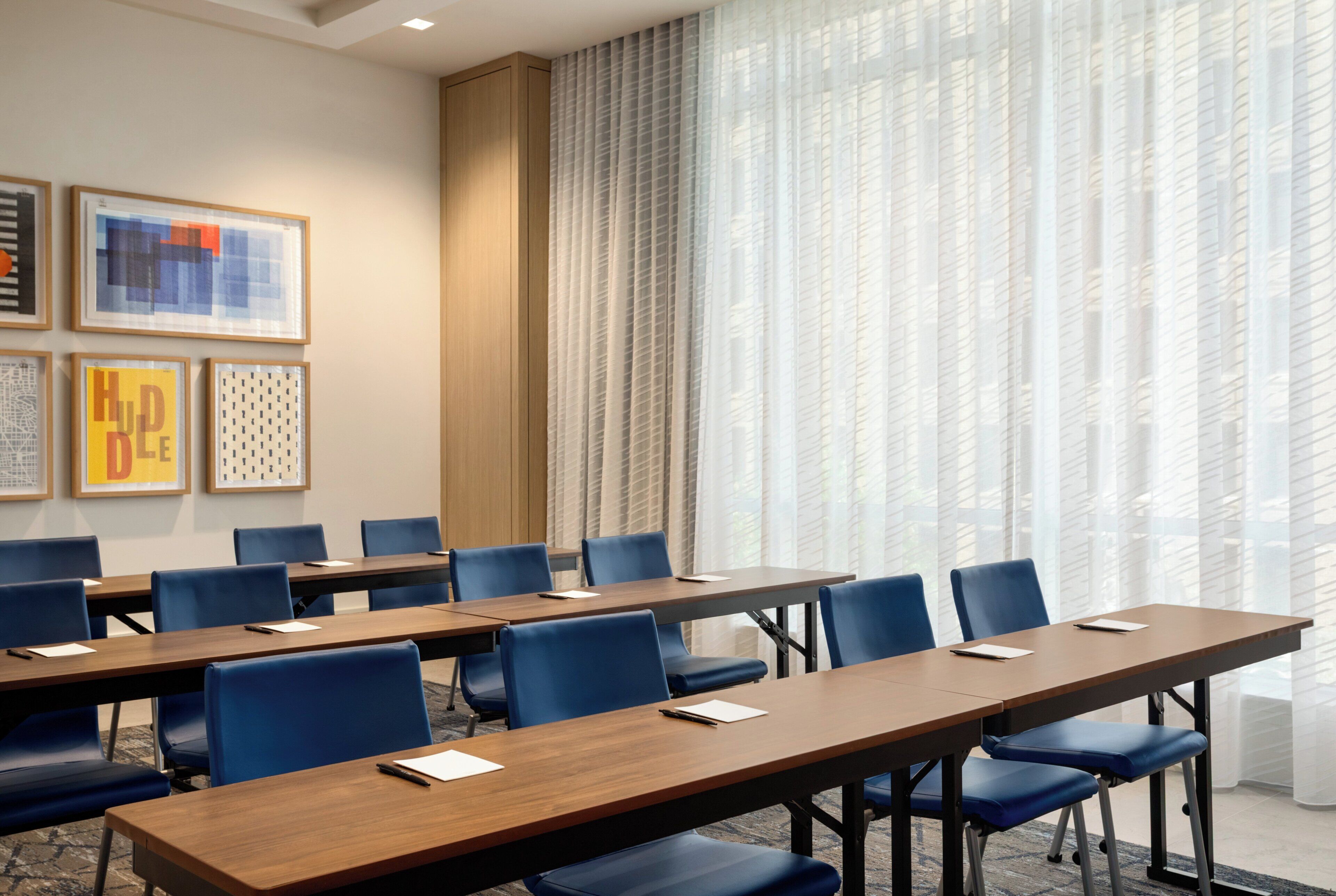 A conference room with long tables and blue chairs, framed artwork on the wall, and sheer curtains on the windows.