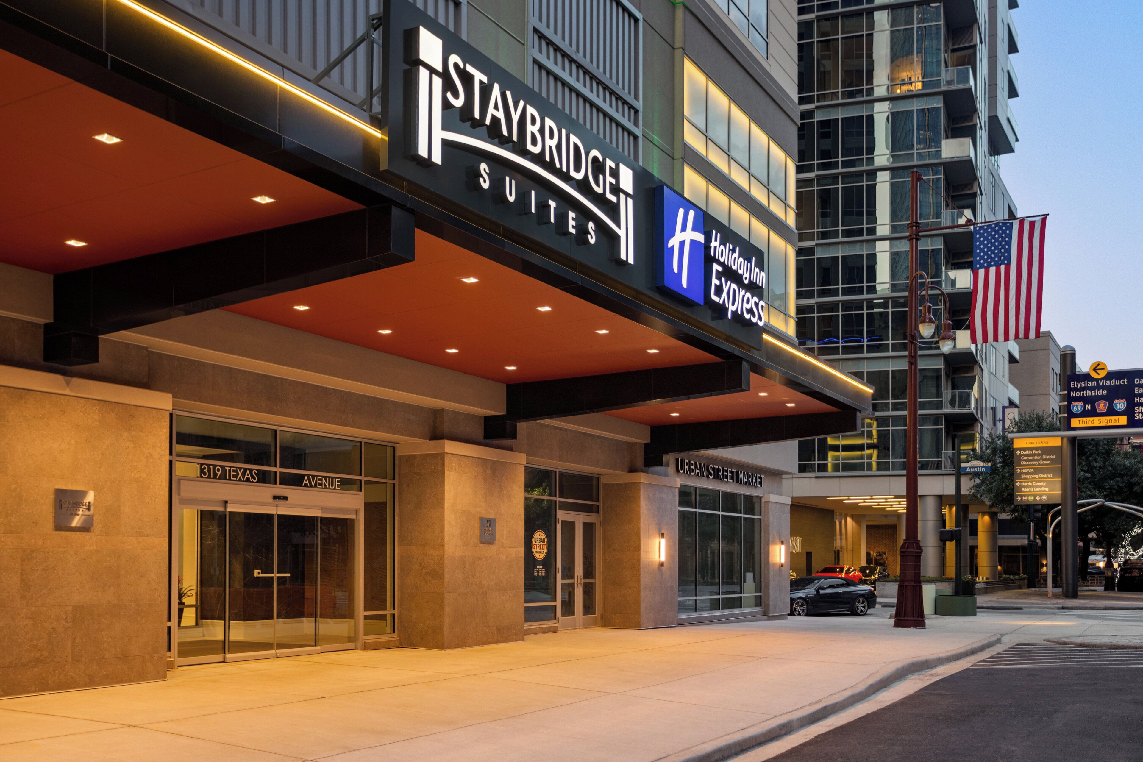 A Staybridge Suites hotel with a glass facade, a sign for Holiday Inn Express, and an American flag.
