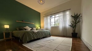 Ein Schlafzimmer mit Bett, Nachttischlampen, einem großen Fenster mit Vorhängen, einer Zimmerpflanze und einem Holzboden.
