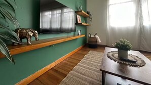 Ein Wohnzimmer mit grüner Wand, einem Wand-Fernseher, Holzregalen, einer kleinen Elefantfigur und einem Fenster mit transparenten Vorhängen.
