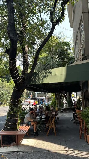 An outdoor seating area with tables and benches under a green awning, surrounded by trees and a building.