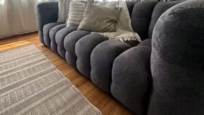 Ein modernes Wohnzimmer mit einem dunkelgrauen Sofa, einem gemusterten Teppich und einem großen Fenster mit transparenten Vorhängen.