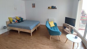 A modern bedroom with a wooden bed, a blue armchair, a small white table, a TV, and a large window. - Large Studio in quiet inner-city location (Dinan)