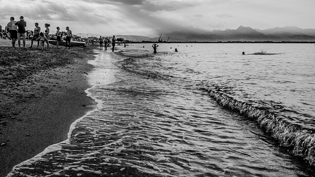 Una playa con gente caminando por la orilla, un mar tranquilo y montañas lejanas bajo un cielo nublado.