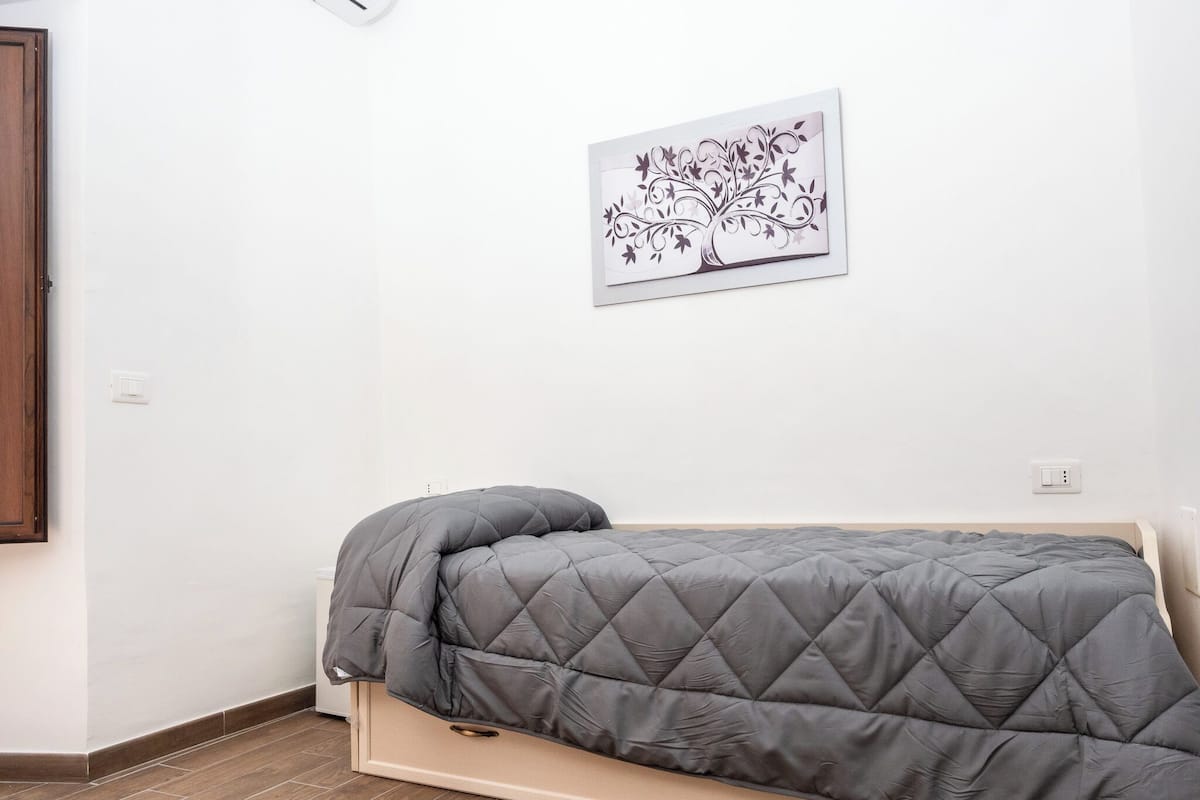 Una cama individual con un colchón gris acolchado y una almohada a juego, ubicada frente a una pared blanca con una pequeña obra de arte floral enmarcada encima.