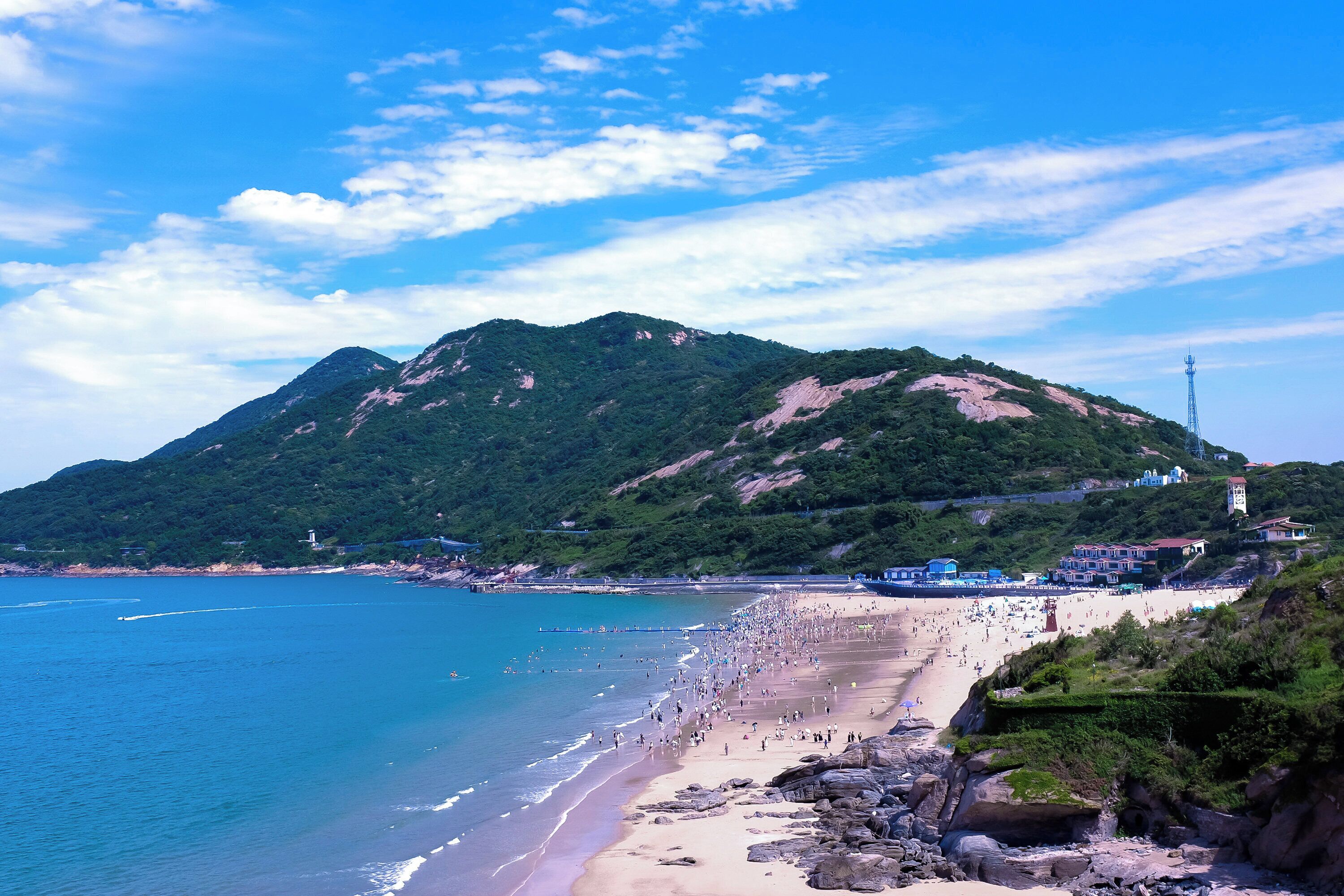 A beach with a mountain in the background, a pier extending into the water, and buildings along the shore.