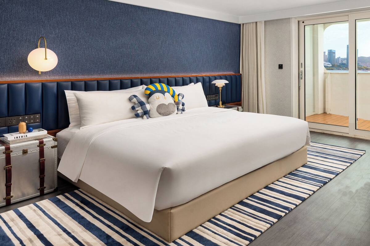 A hotel room with a large bed, a blue headboard, a striped rug, and a suitcase on the nightstand.