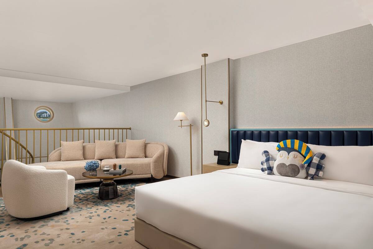A hotel room with a bed, a sofa, a chair, a coffee table, a lamp, and a wall-mounted phone.
