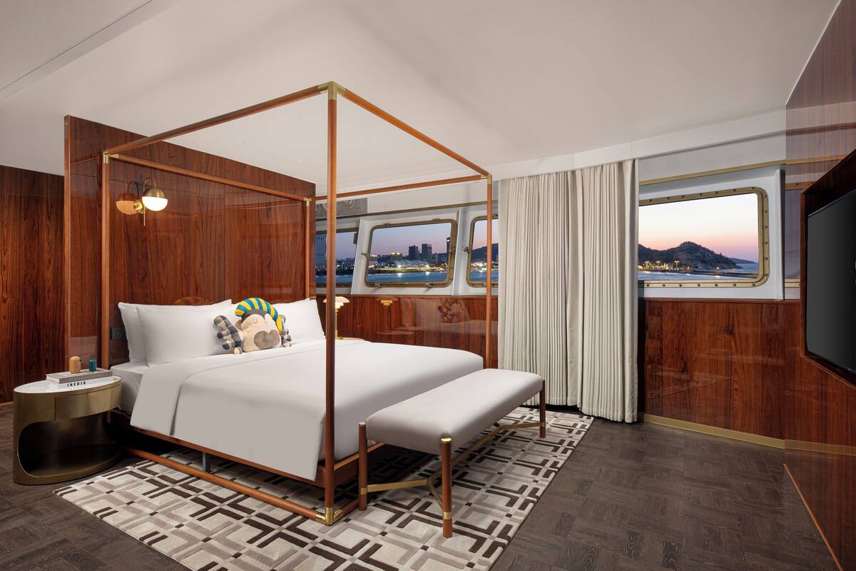 A four-poster bed with a city view, a bedside table with a lamp, and a large window with curtains.