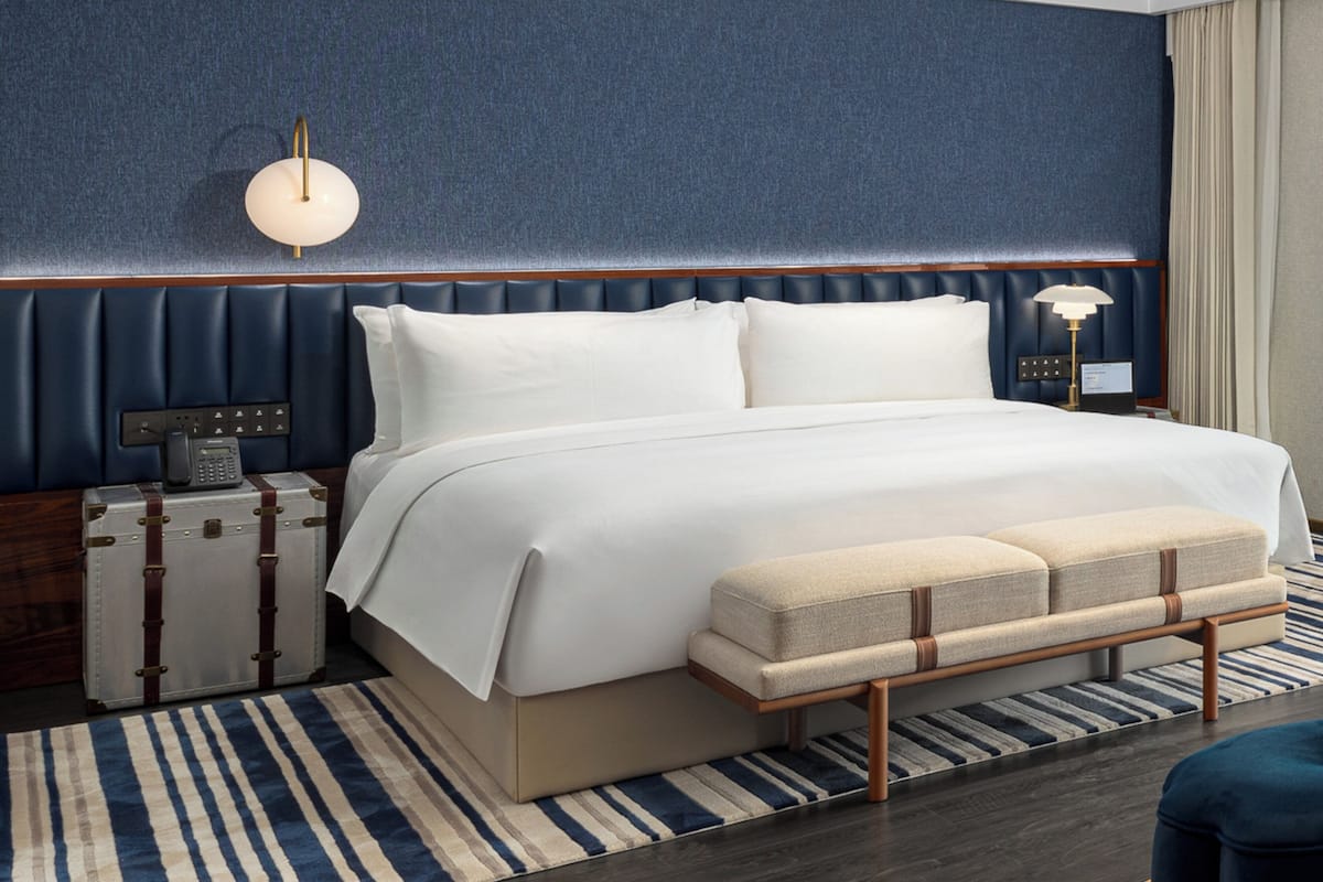 A hotel room with a large bed, a bedside table with a lamp, a suitcase, and a striped rug.