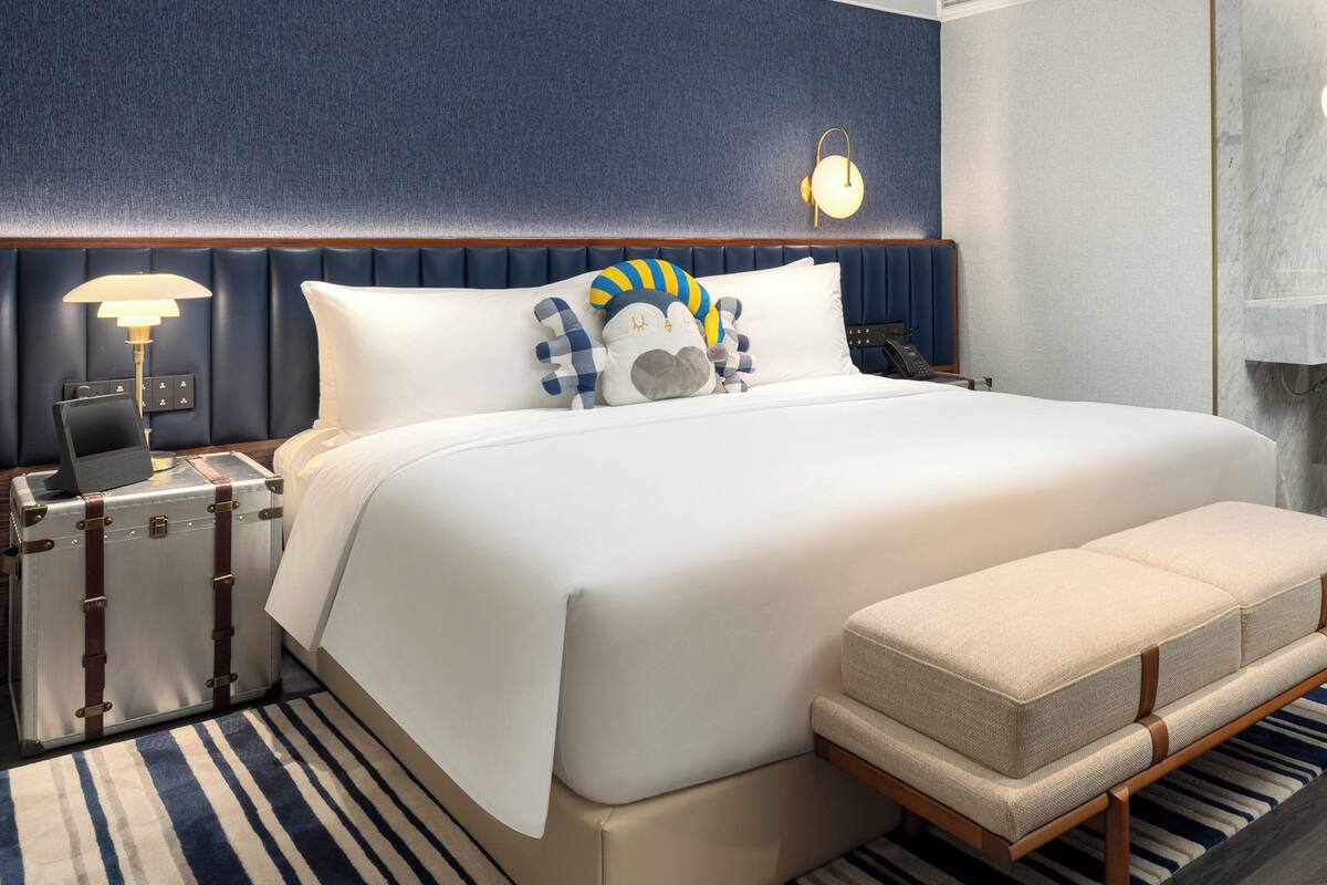 A hotel room with a large bed, a bedside table with a lamp, a suitcase, and a bench.