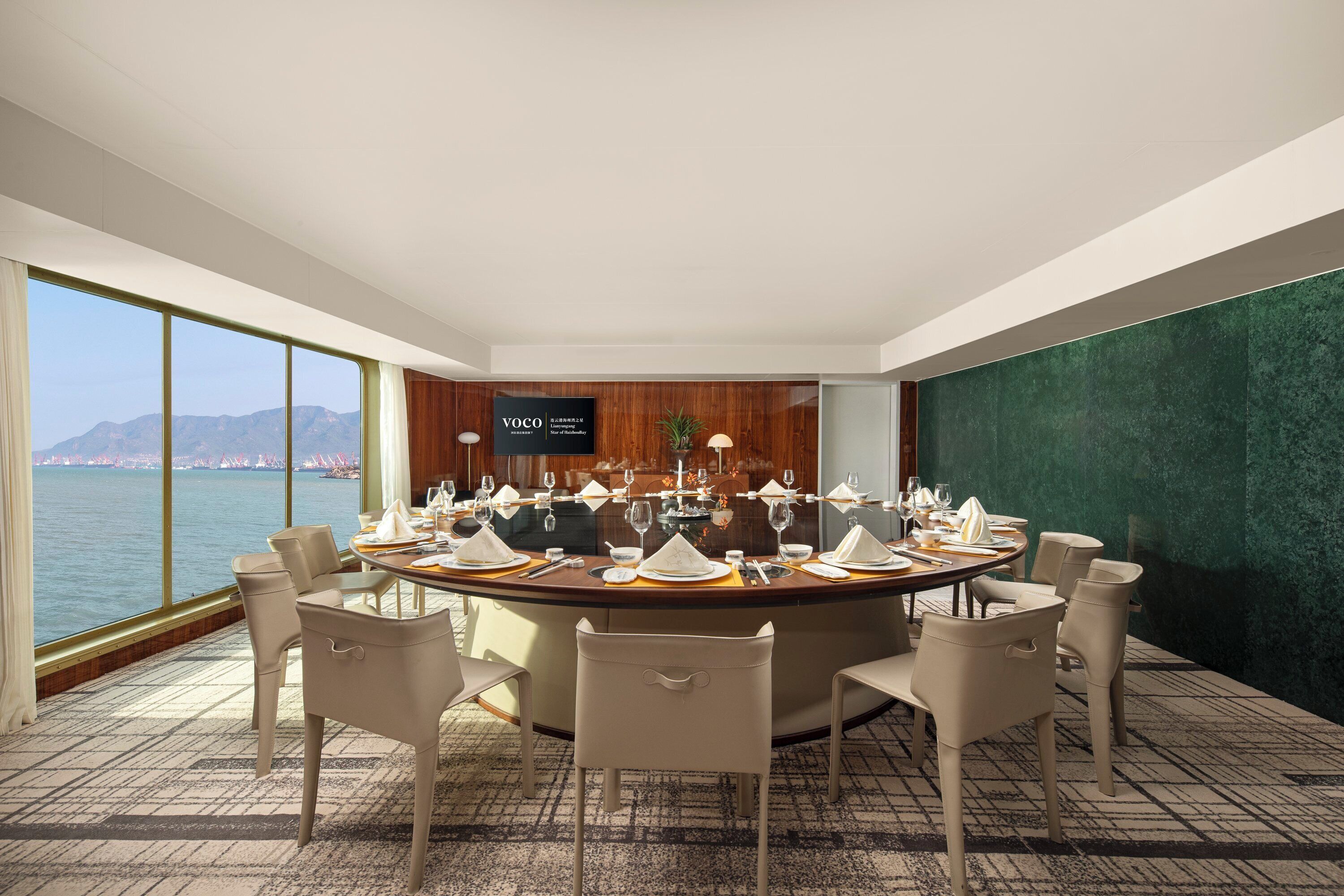 Un salón de conferencias con una mesa redonda lista para una reunión, ventanas amplias con vista a un cuerpo de agua y montañas distantes, y una pared verde con textura.
