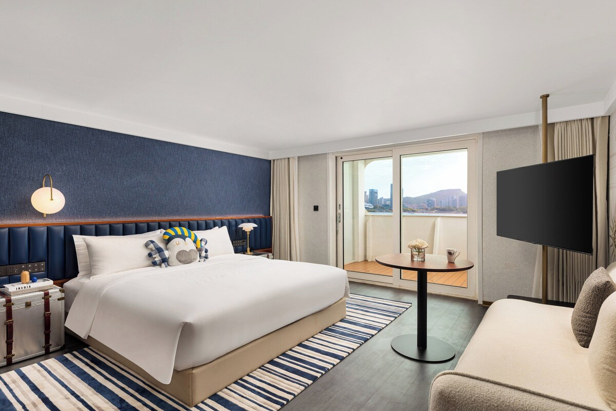 A hotel room with a large bed, a TV, a desk, and a balcony with a city view.