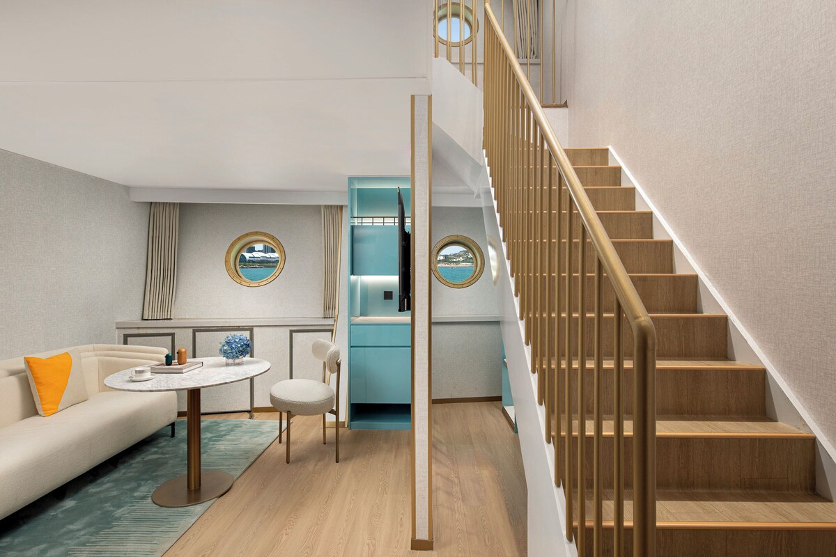 A modern hotel room with a wooden staircase, a round mirror, a sofa, a small table, and a view of the sea through a round window.