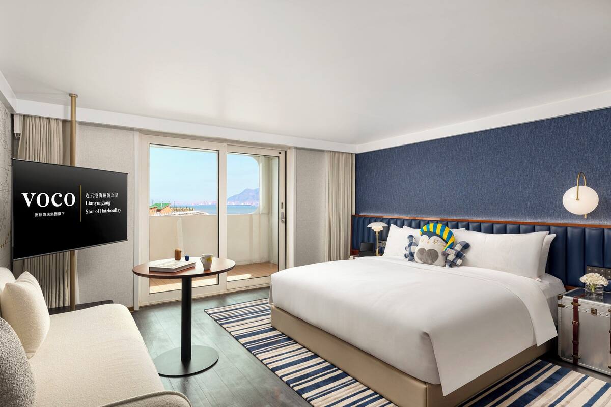 A hotel room with a large bed, a TV, a balcony with a view, and a small table with a lamp.