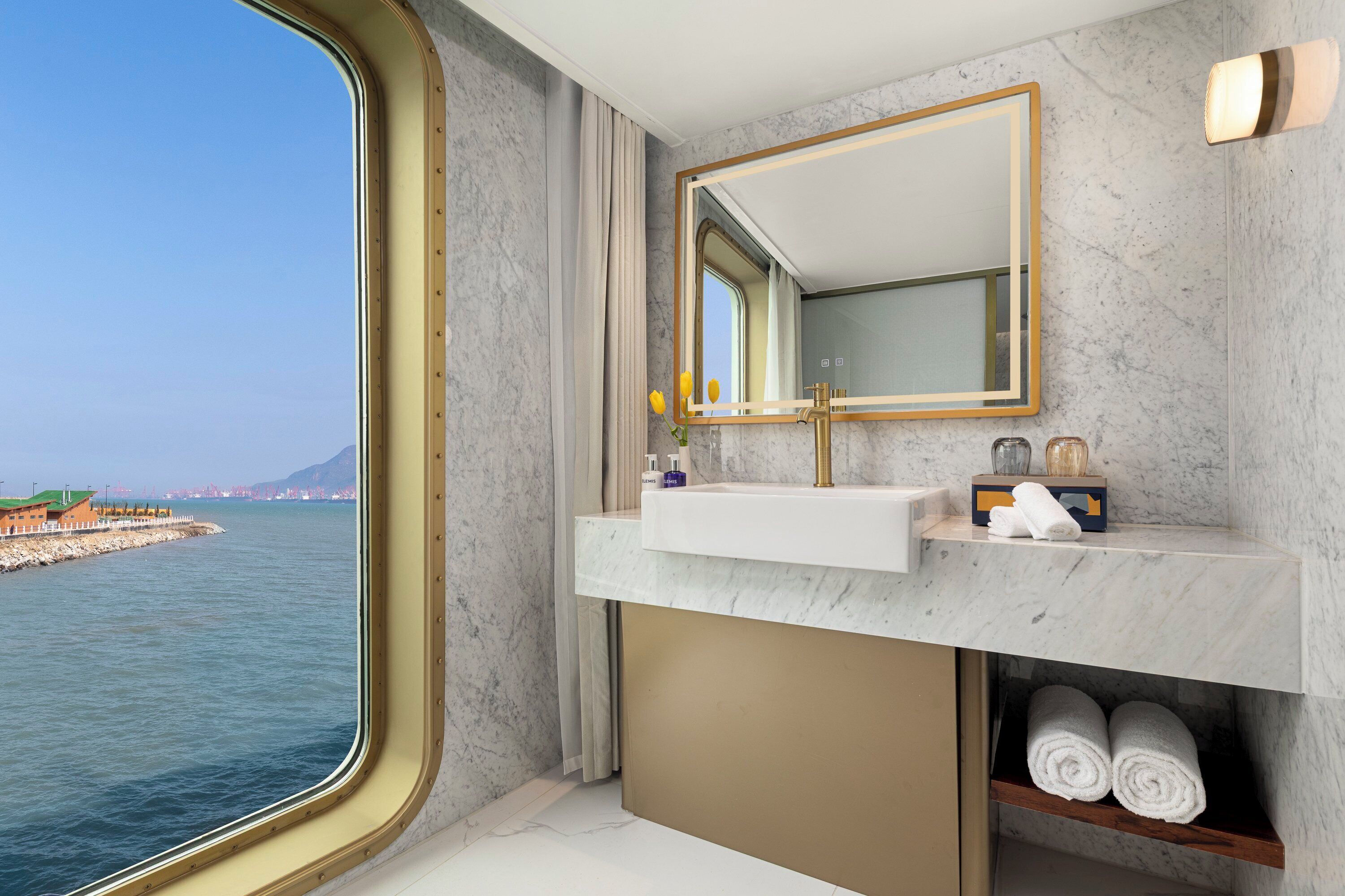 Een badkamer met een marmeren wastafel, een spiegel met gouden lijst en uitzicht op zee.