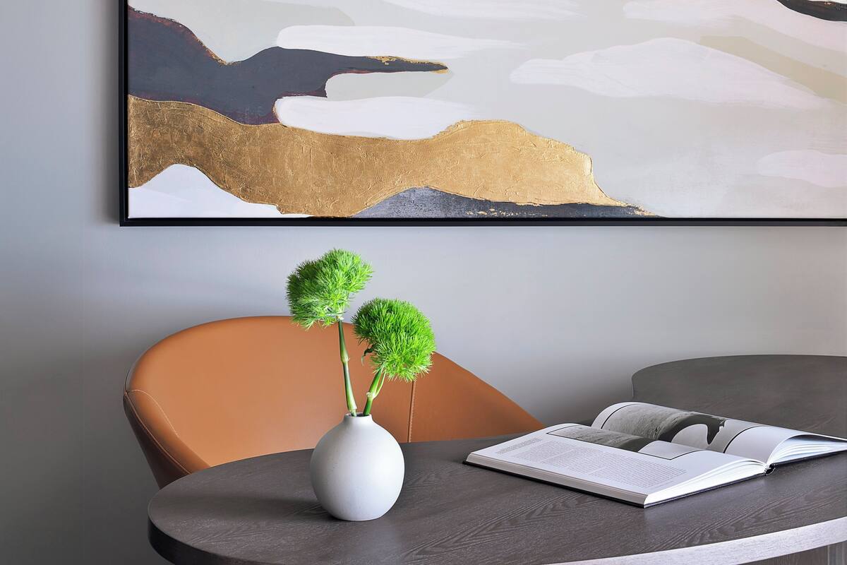 A modern room with a round table, a white vase with green plants, a brown chair, and a large abstract painting on the wall.