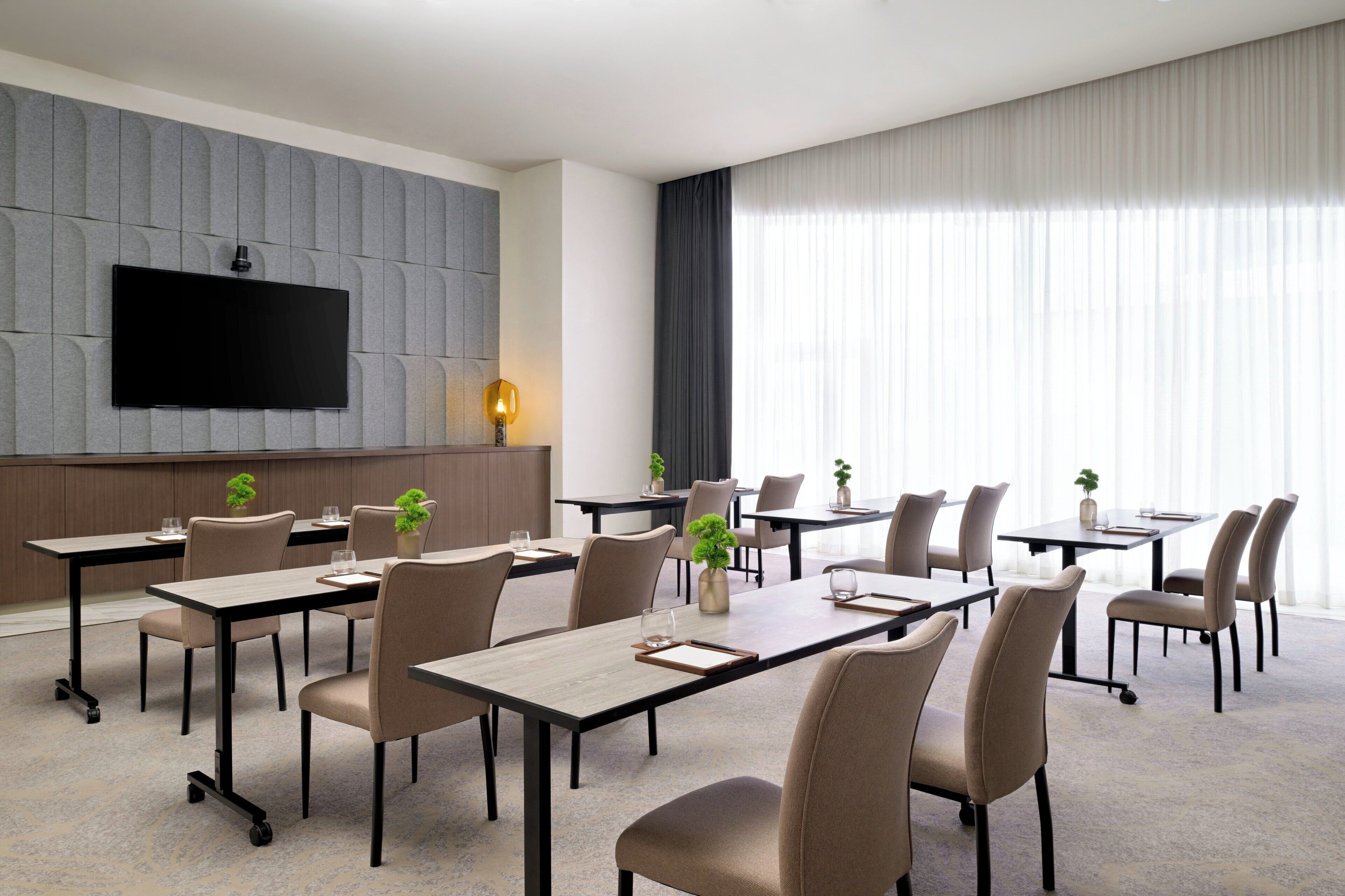 A modern hotel conference room with multiple tables, chairs, a flat-screen TV, and large windows with sheer curtains.