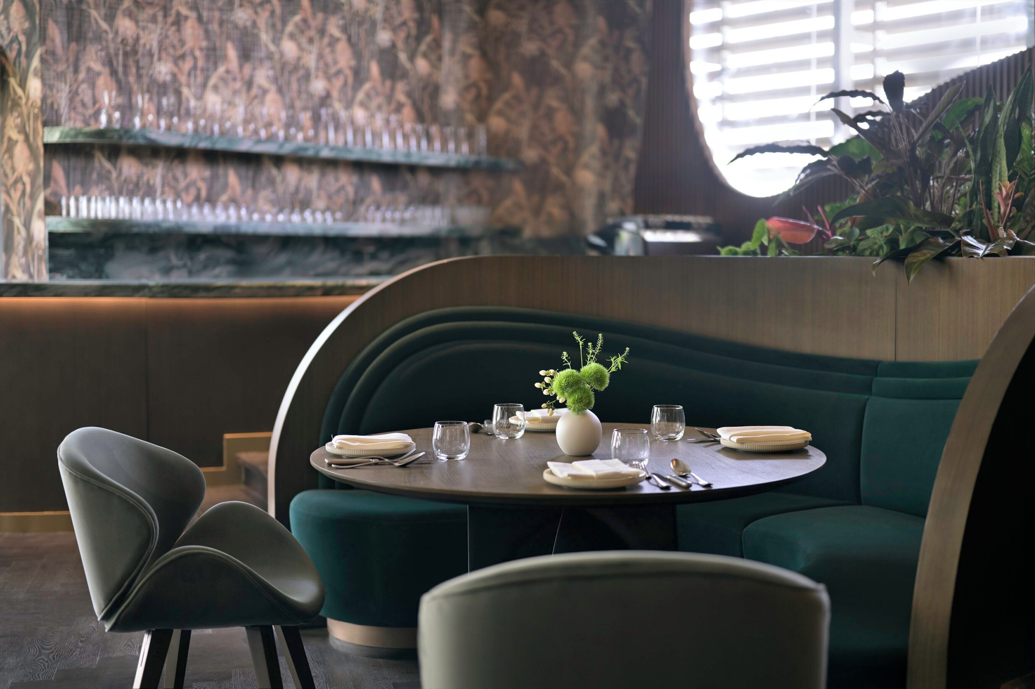 A dining area with a round table set for two, a green upholstered booth, and a decorative vase with greenery.