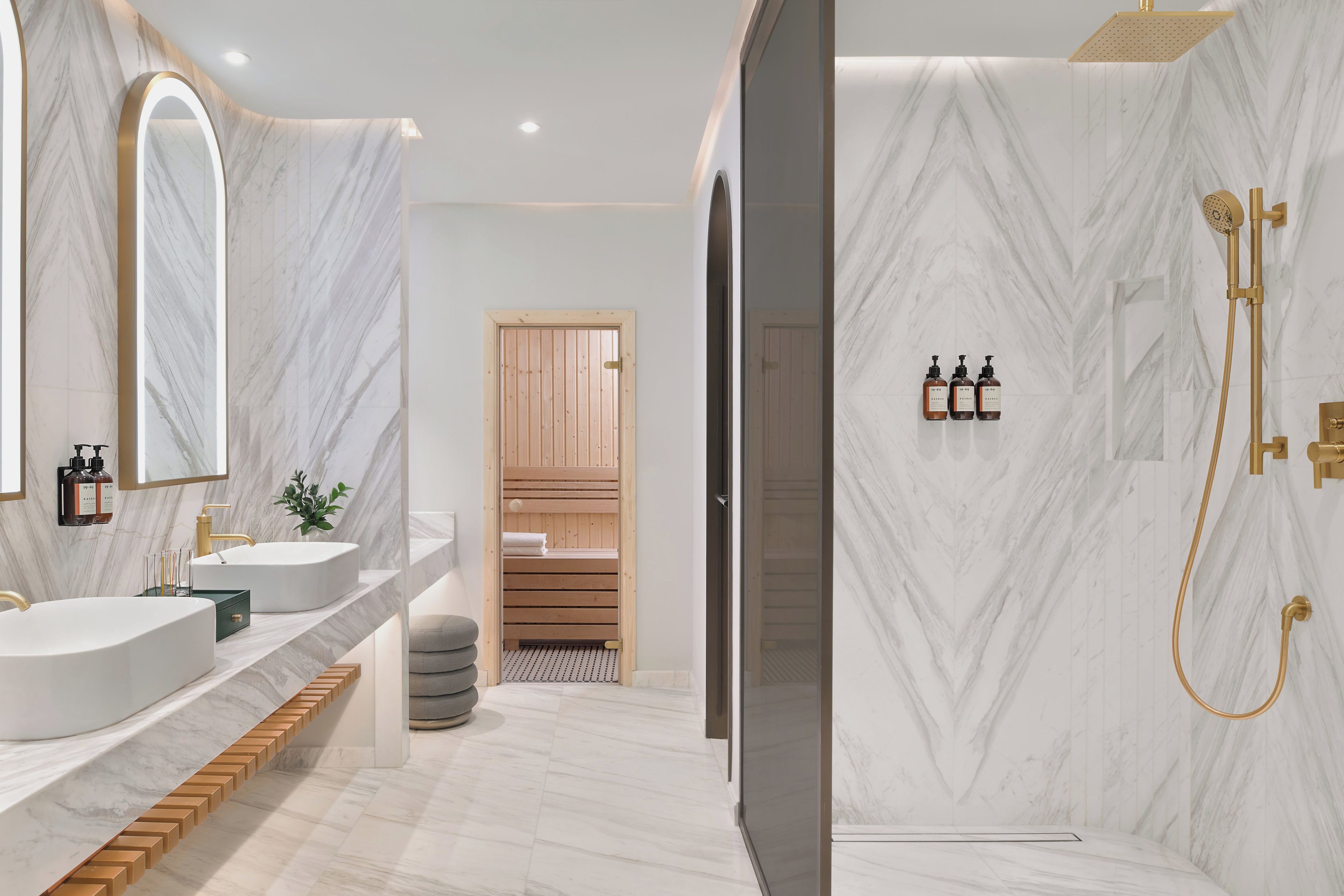 Ein modernes Badezimmer mit zwei Waschbecken, einem großen Spiegel, einer Dusche mit goldener Armatur und einer hölzernen Sitzbank.