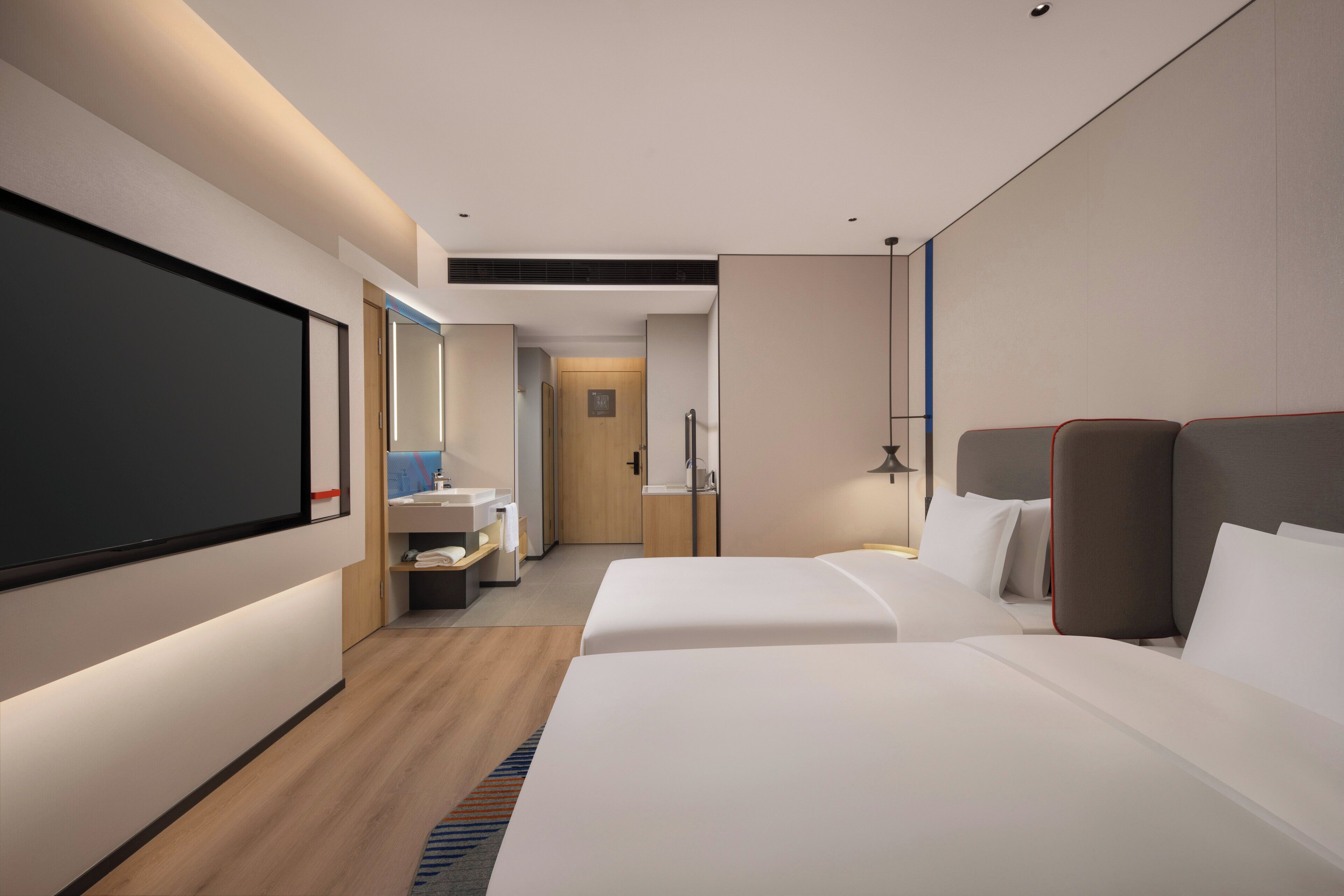 A modern hotel room with two beds, a large flat-screen TV, a bathroom with a sink and mirror, and a wooden floor.