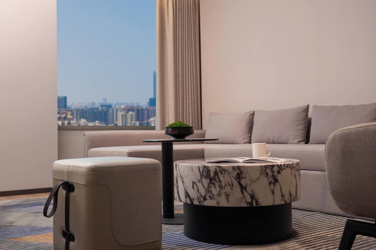 A modern hotel room with a city view, a sofa, a marble coffee table, and a suitcase.