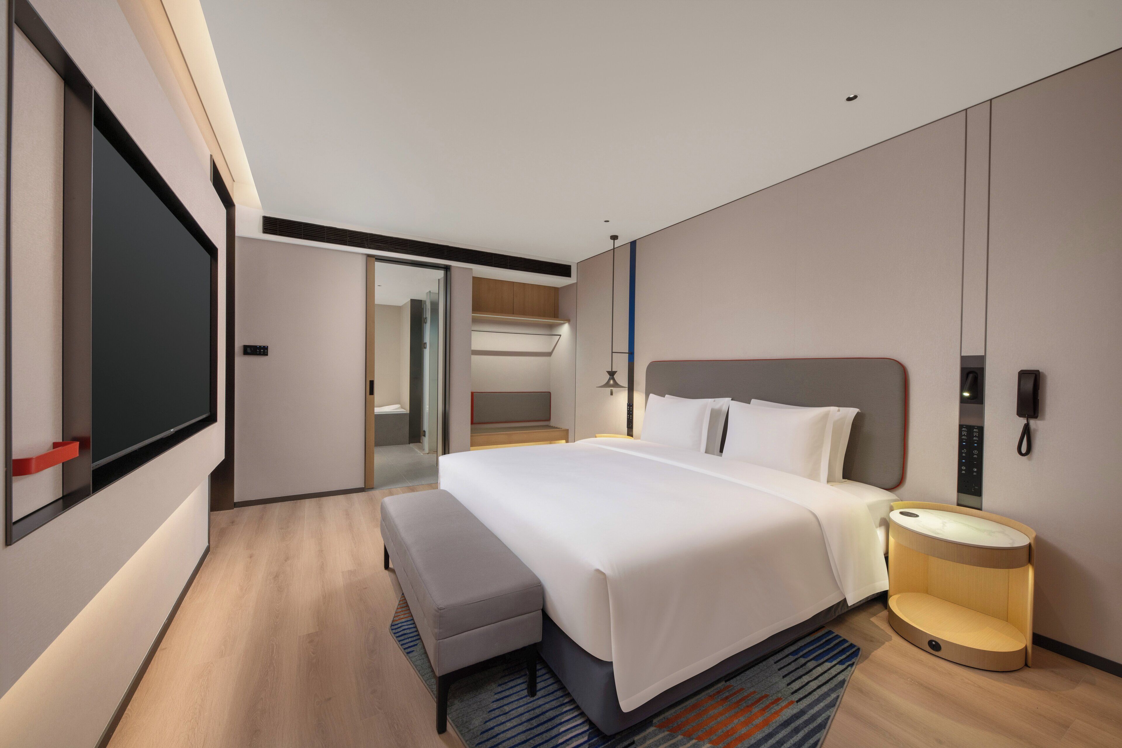 A modern hotel room with a large bed, a bedside table with a lamp, a bench, a TV, and a wardrobe.