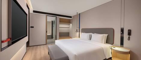 A modern hotel room with a large bed, a bedside table with a lamp, a bench, a TV, and a wardrobe.