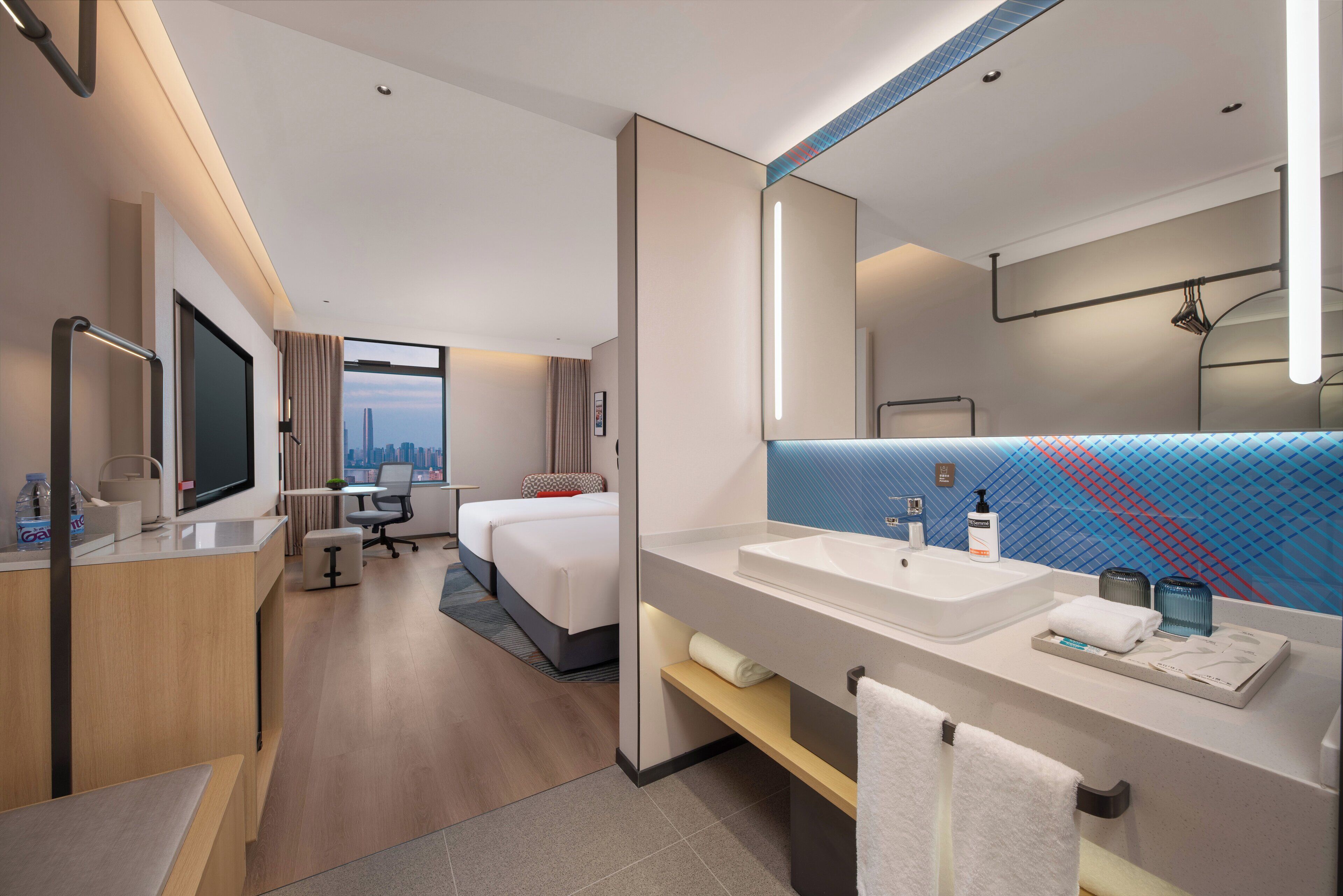 A hotel room with a large bed, a desk with a computer, a bathroom with a sink and mirror, and a city view.