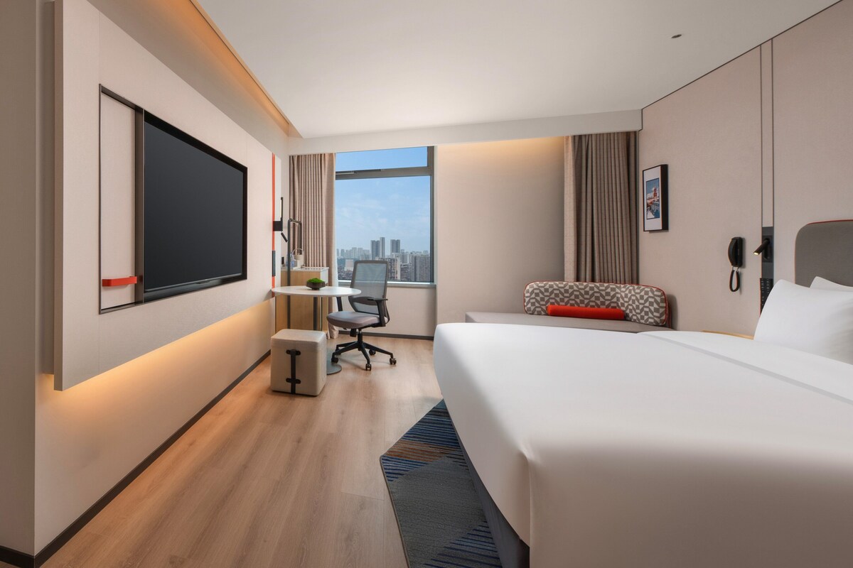 Een moderne hotelkamer met een groot bed, een bureau met stoel, een flatscreen televisie, een telefoon en een raam met uitzicht op de stad.