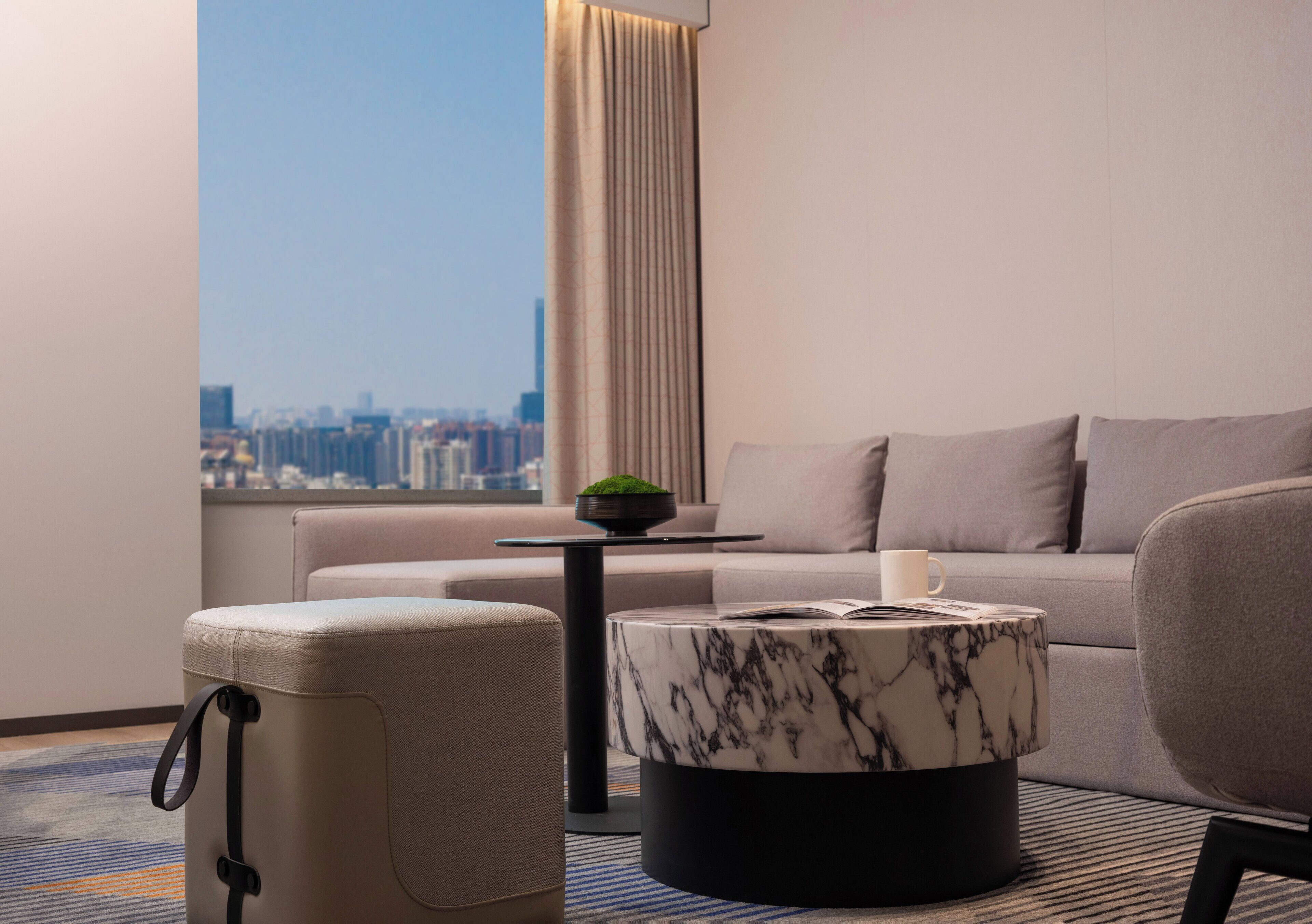 Ein modernes Hotelzimmer mit Blick auf die Stadt, einer Couch, einem Marmortisch und einem Koffer.