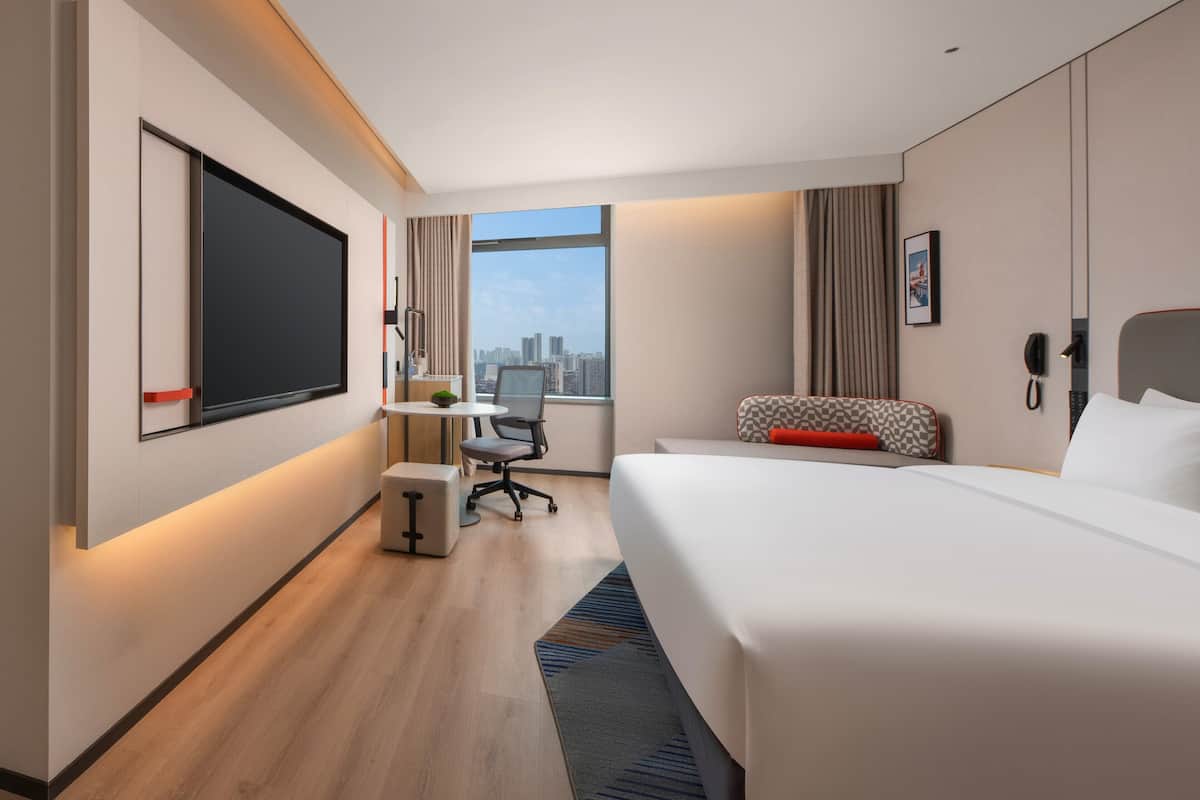 A modern hotel room with a large bed, a desk with a chair, a flat-screen TV, a phone, and a window with a city view.