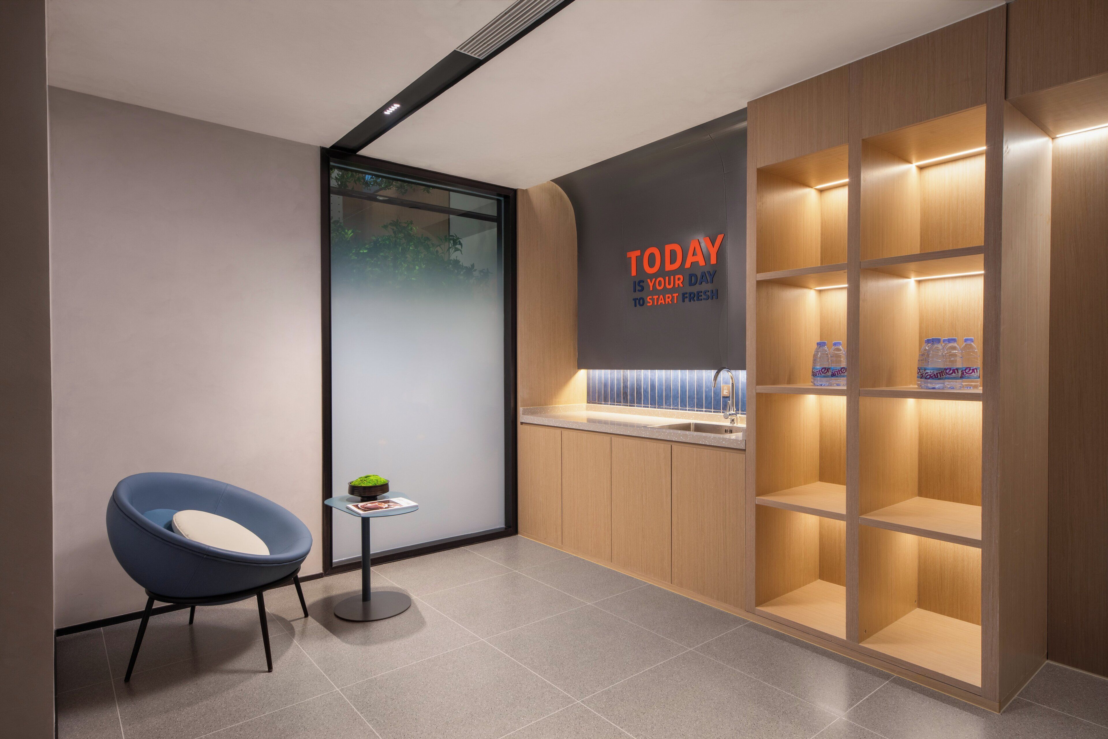 A modern reception area with a blue chair, a small table with a plant, and a wooden shelving unit with water bottles.