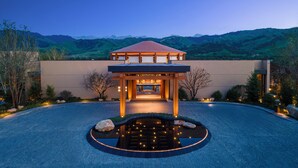 A modern building with a red roof, a covered entrance, and a circular water feature in front. - Hotel Indigo Nalati by IHG (Xinyuan)