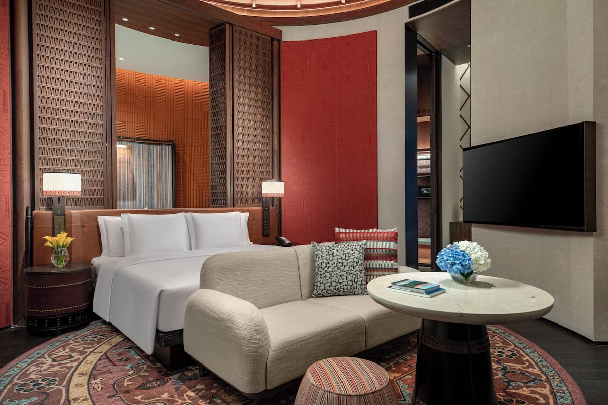 Habitación de hotel con cama, sofá, mesa redonda, televisor y alfombra estampada.