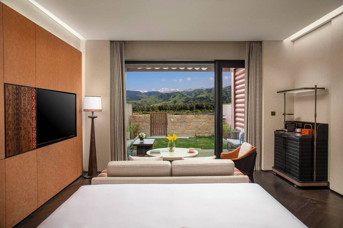 Habitación de hotel con una cama grande, un televisor de pantalla plana, un sofá, una mesa de comedor con un florero y vistas a las montañas a través de una puerta corrediza.