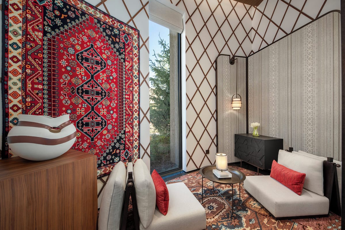 A modern living room with a sofa, armchair, and a patterned rug. There is a wooden cabinet and a black side table with a lamp.