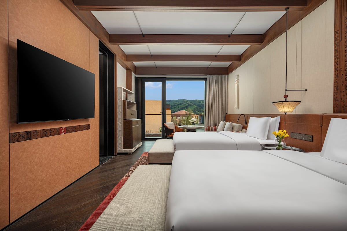 Habitación de hotel con dos camas, televisión, balcón con vistas y zona de comedor.