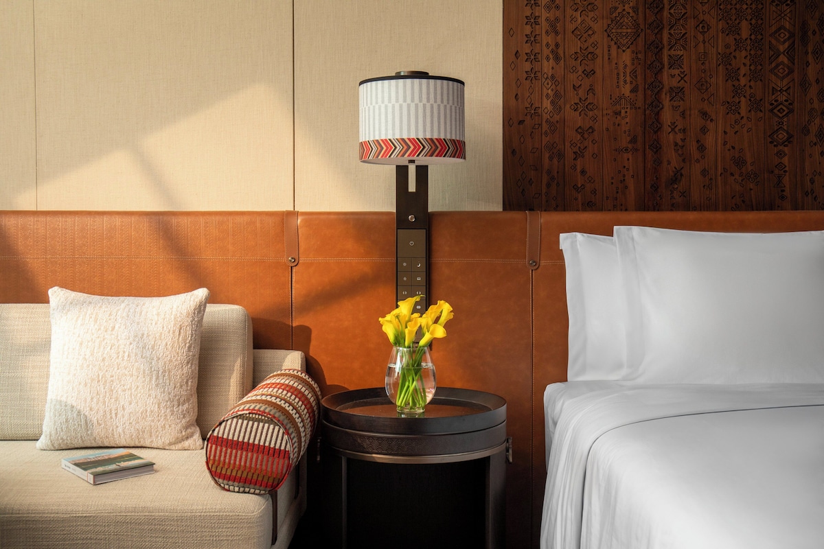 Una habitación de hotel con cama, sofá, mesita de noche con lámpara y un jarrón con flores.