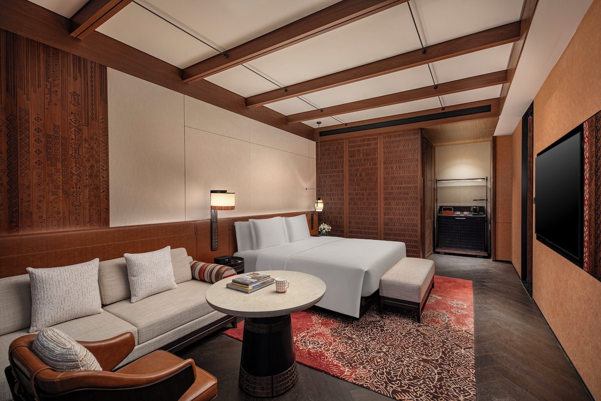 Una habitación de hotel moderna con cama, sofá, sillón, una mesita y televisor.