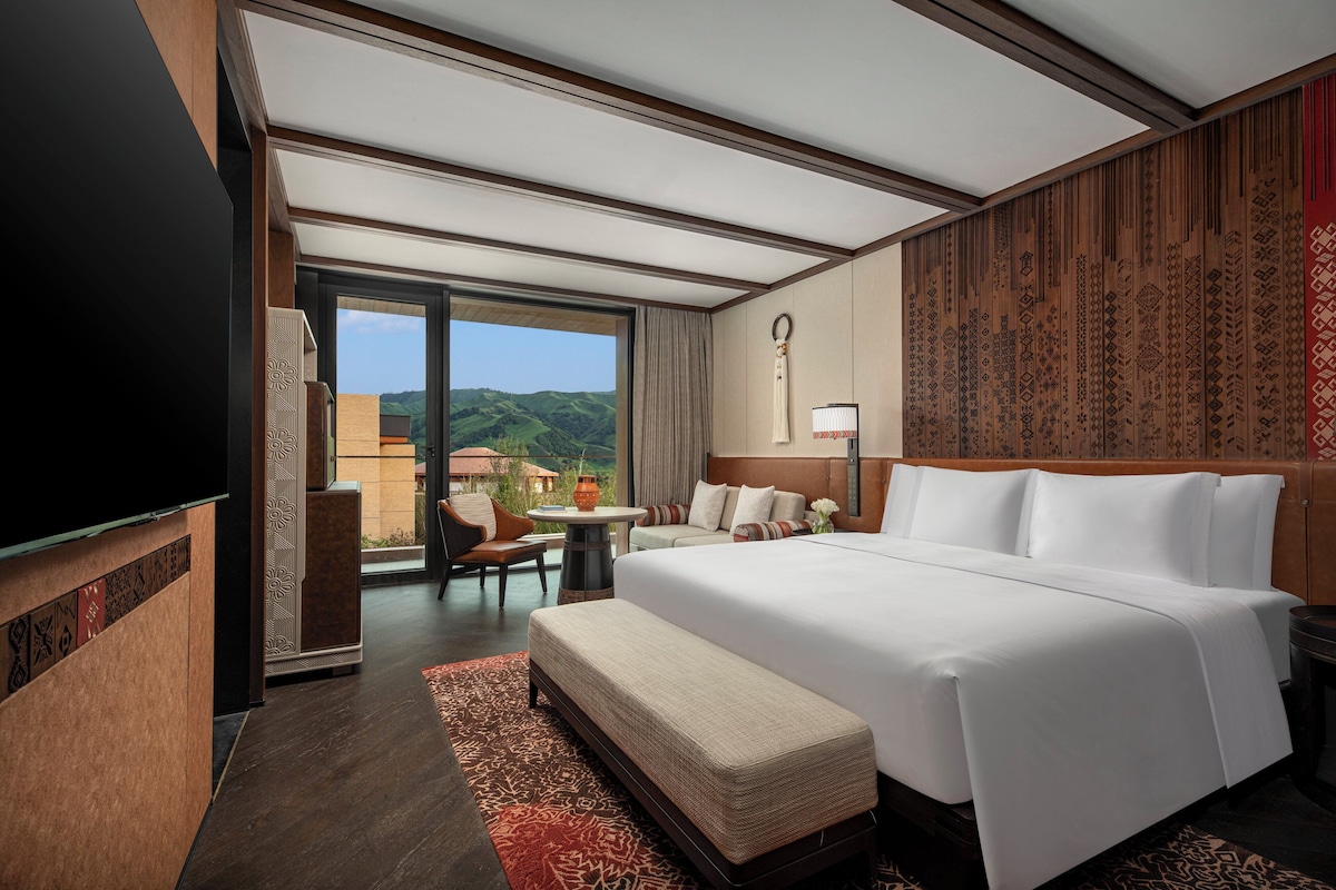 Habitación de hotel con una cama grande, un televisor de pantalla plana, una mesita y un espacio de descanso con vistas a las colinas.