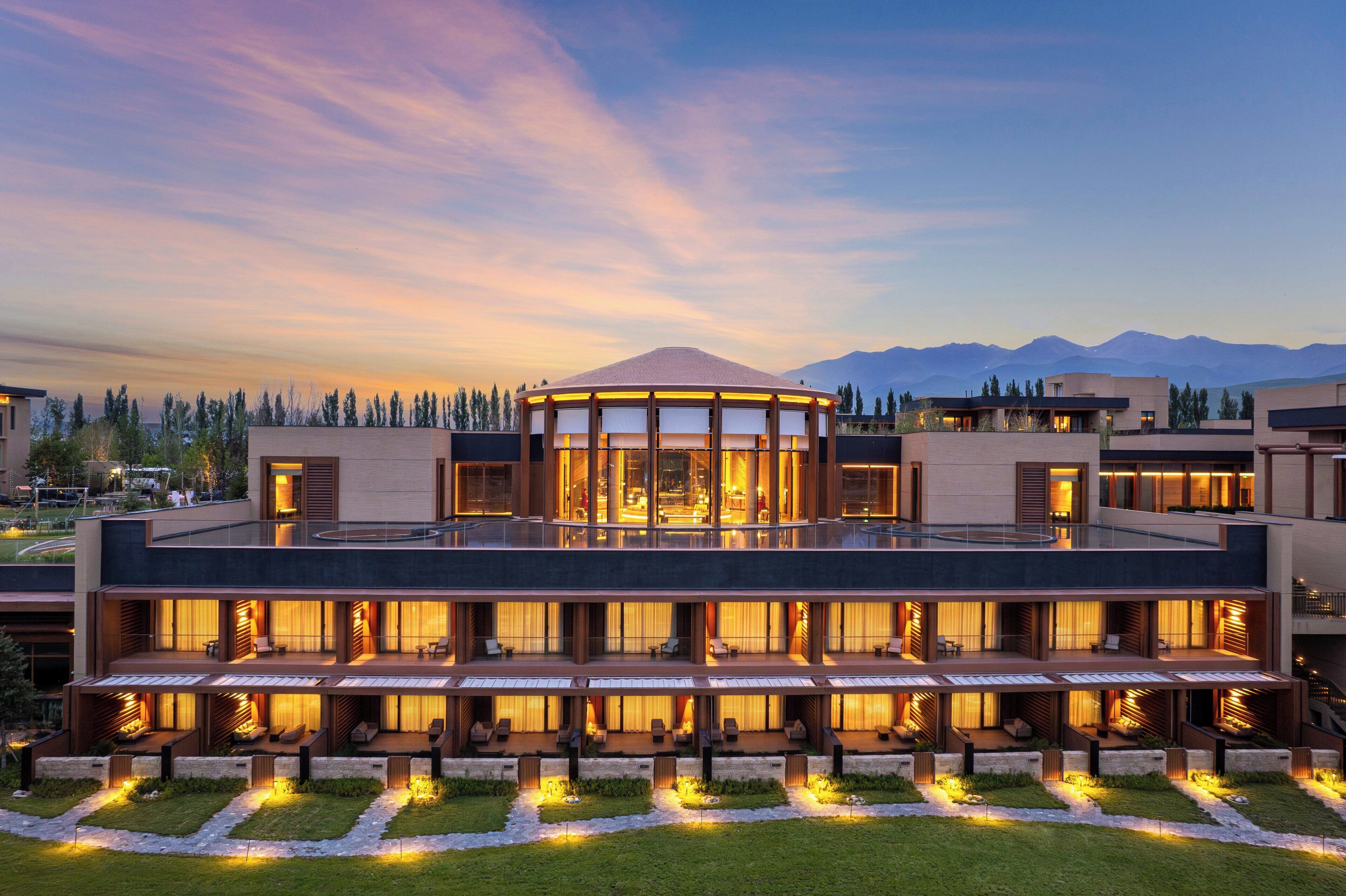 Un hotel moderno con una estructura central de vidrio, varios balcones y vistas a montañas.