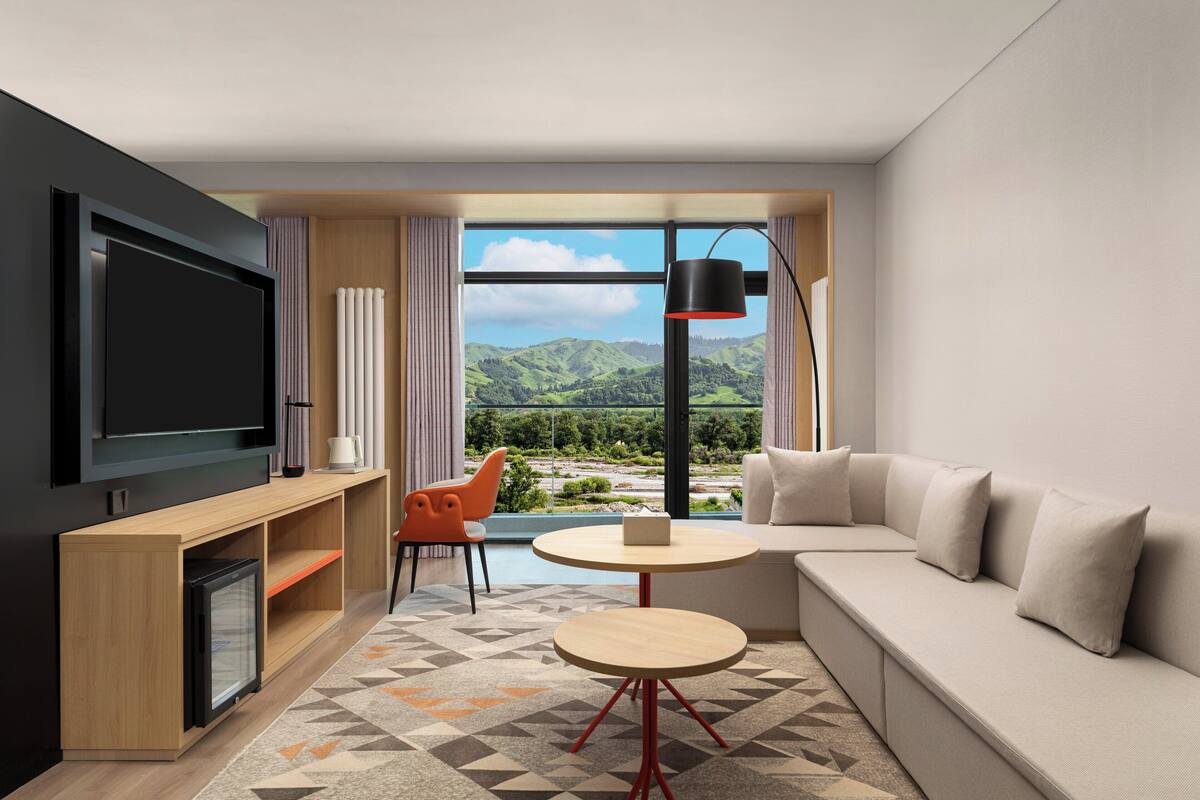 A modern living room with a large TV, a sofa, a small table, and a chair. There is a window with a view of green hills.