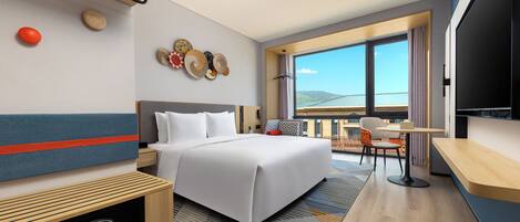 A modern hotel room with a large bed, a desk, a television, and a balcony with a view.